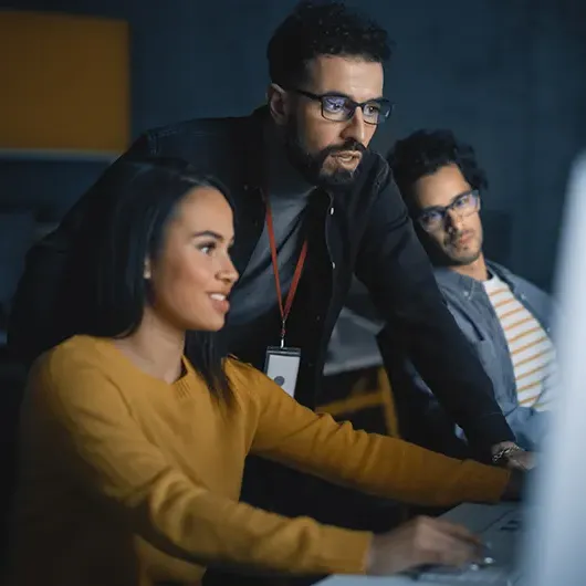  Three software developers talking about programming issue leaned over desk looking at computer monitor